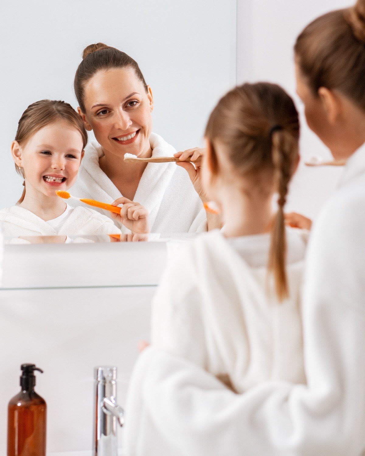 A mother and her daughter brushing their teeth together at the bathroom mirror — the daily routine that preventive dentistry is built on